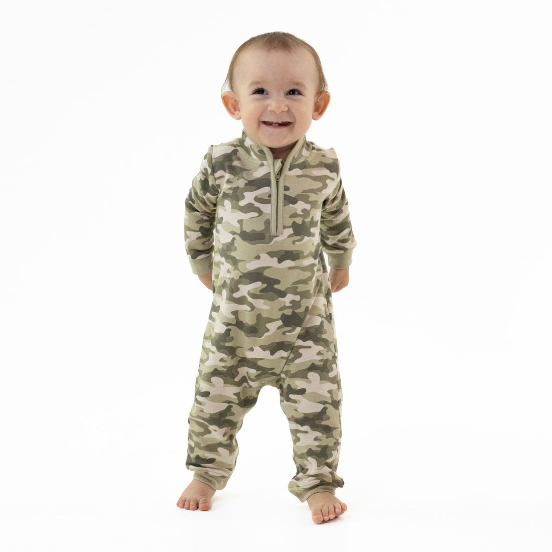 A smiling baby standing on a white background, wearing a long-sleeve camouflage one-piece jumpsuit with a half-zip neckline.