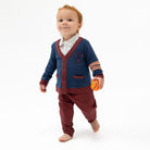 A baby walking barefoot on a white studio background, wearing a navy blue cardigan with burgundy trim over a striped shirt and burgundy pants, while holding a small orange ball.