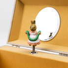 A small rabbit figurine in a white dress with a green wreath stands on a spring-loaded metal handle inside a tan wooden box lid, with a round mirror attached to the inner lid.