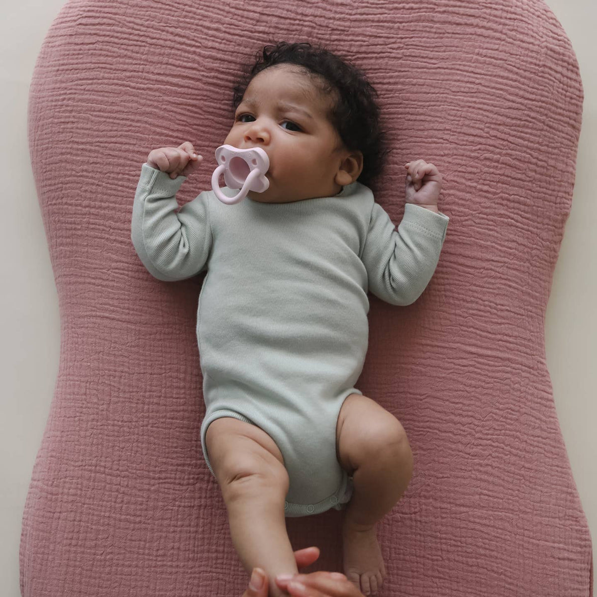 Baby lying on a pink cushion wearing a light green onesie with a pink pacifier.