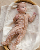 A baby lying on a cream textured blanket inside a woven bassinet, wearing a beige footed one-piece sleeper with a small white floral print and a matching headband.
