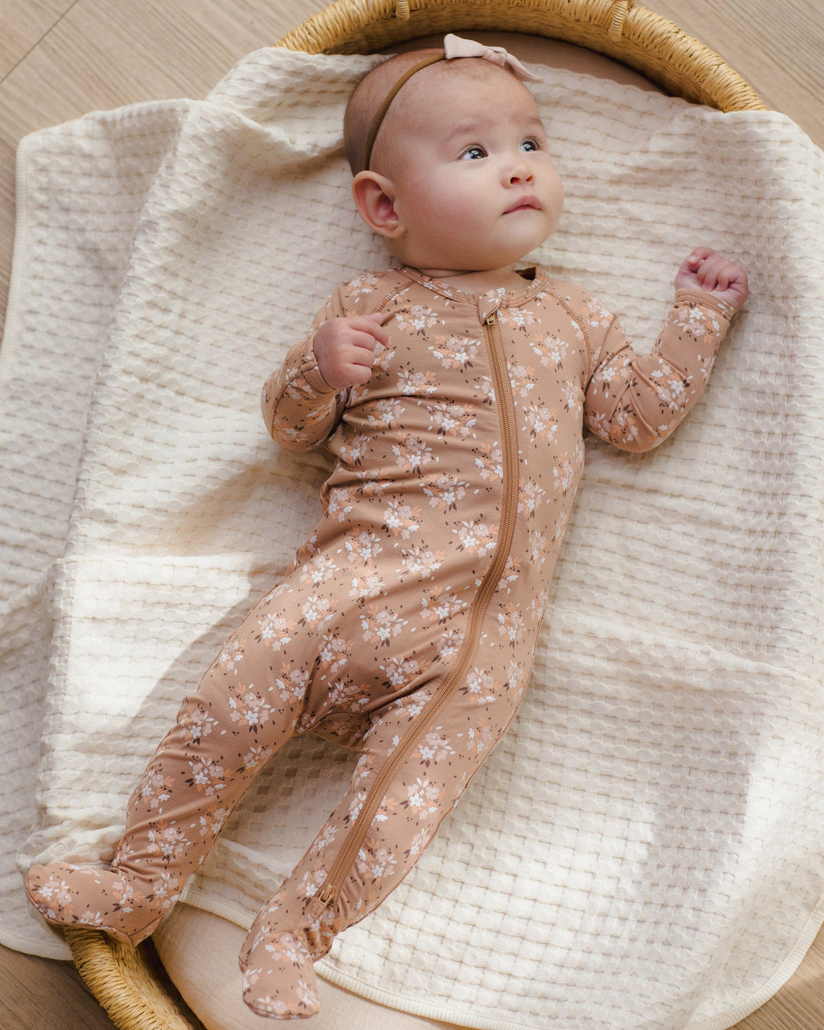 A baby lying on a cream textured blanket inside a woven bassinet, wearing a beige footed one-piece sleeper with a small white floral print and a matching headband.