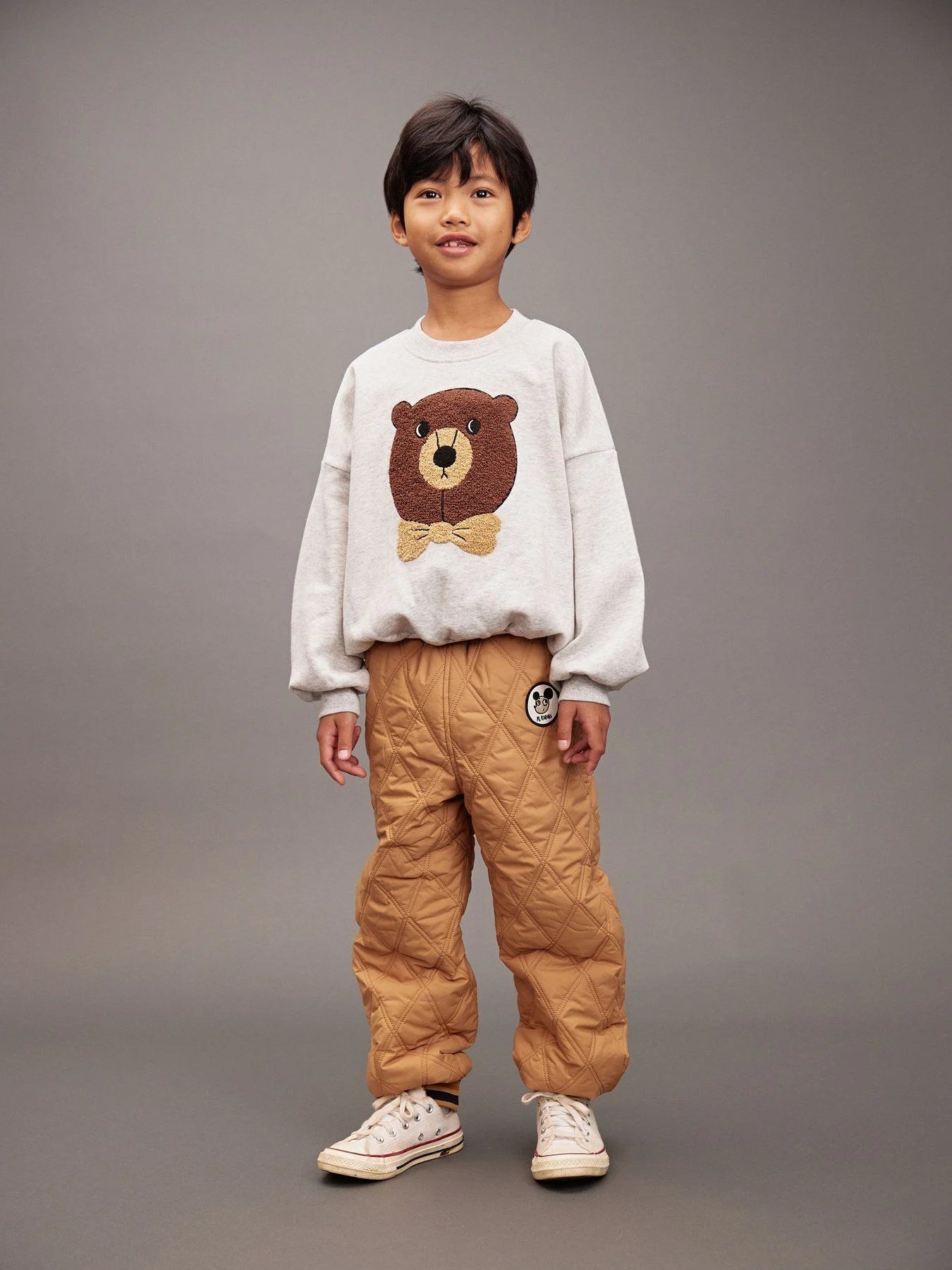 Child wearing a white sweater with a bear design and brown pants on a gray background