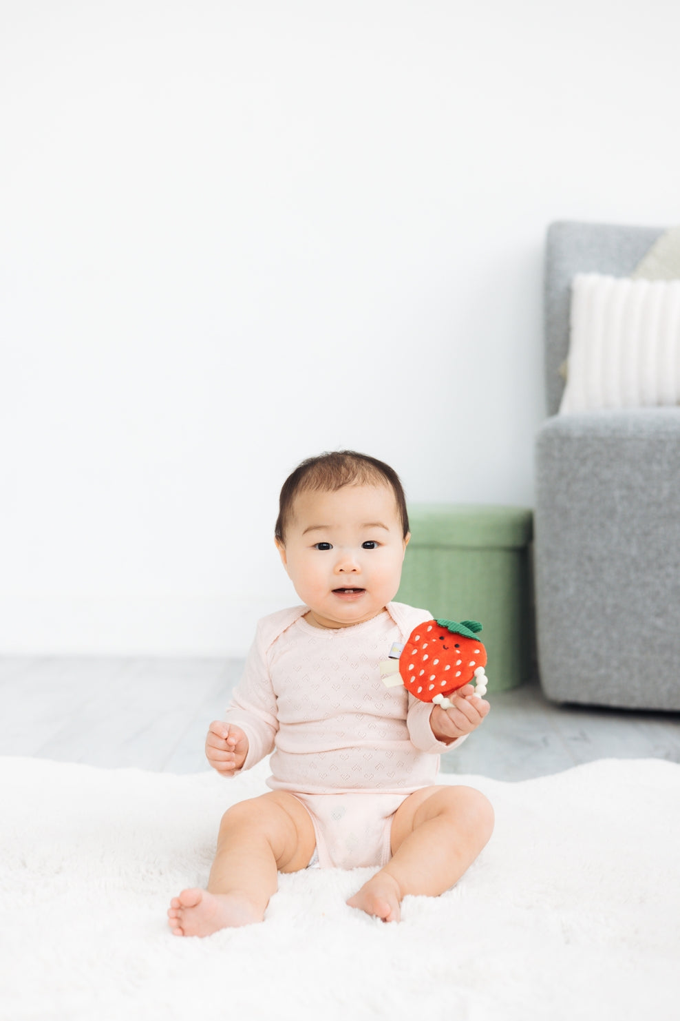 Baby holding a strawberry-shaped toy in a room with a couch and green container.