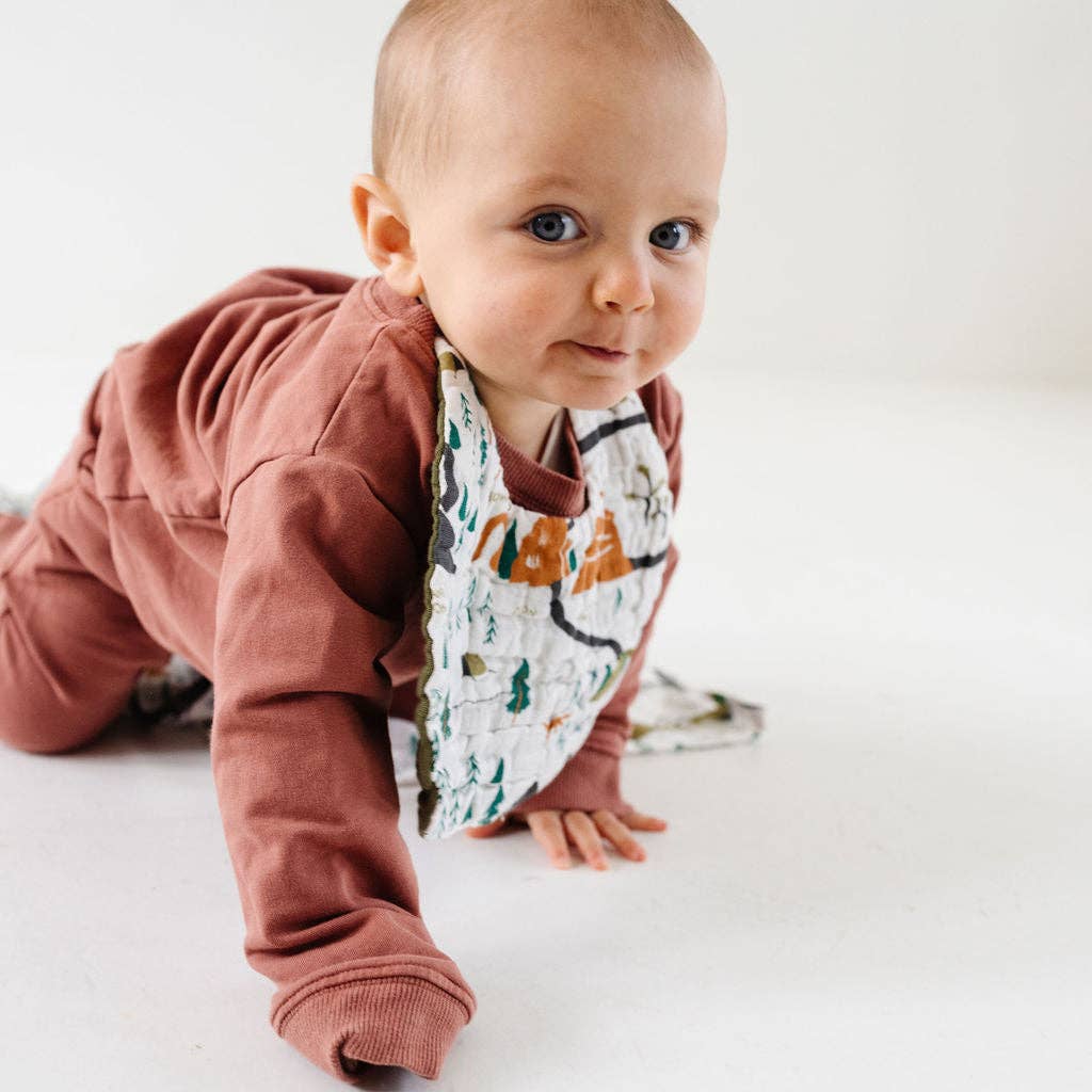 A baby crawling on a white floor wearing a dusty rose long-sleeve romper and a patterned bib with a cartoon character.