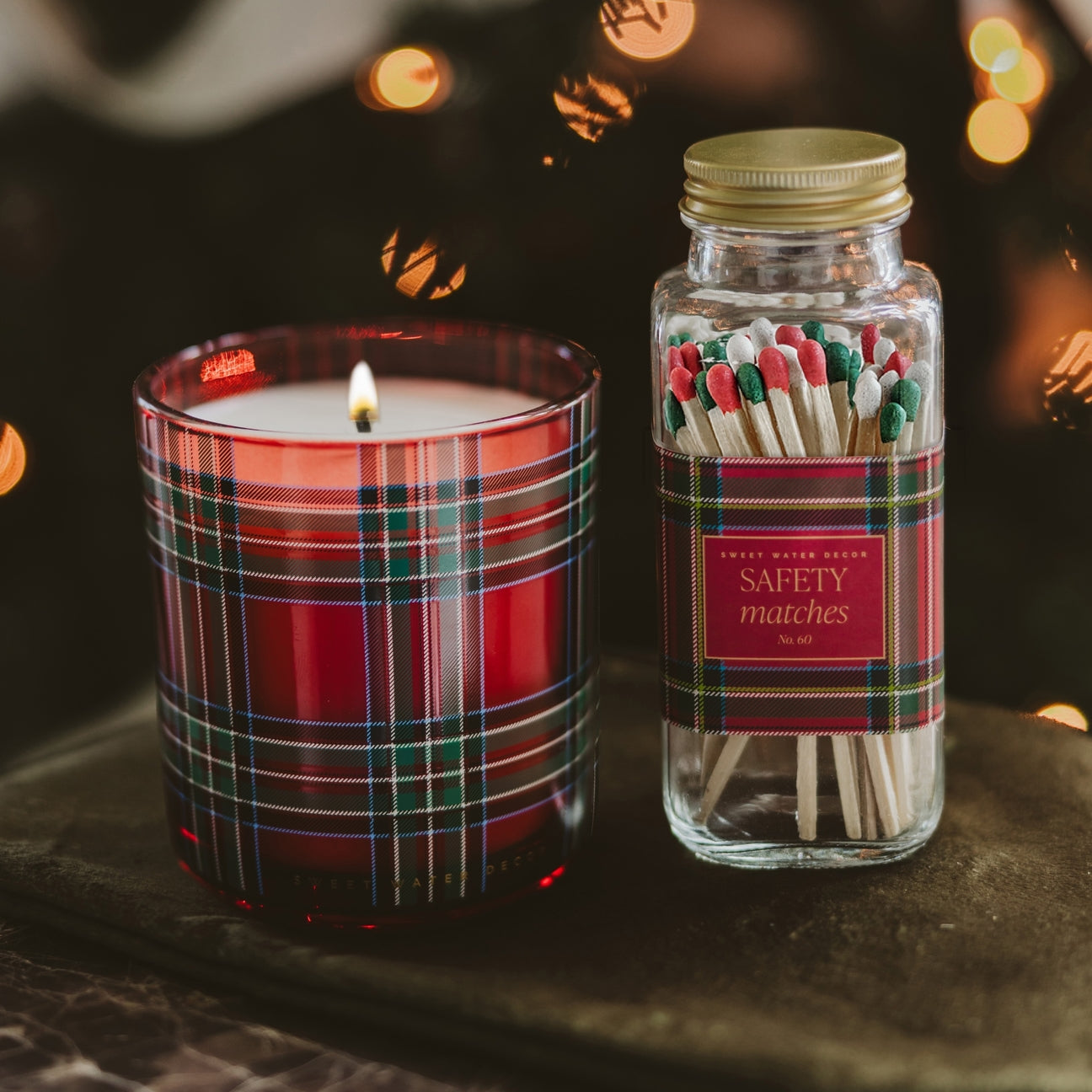 A holiday-themed gift set featuring a lit red plaid scented candle in a glass jar and a matching glass jar of safety matches with a gold lid and red plaid label, set on a dark surface with warm bokeh lights in the background.