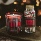 A holiday-themed gift set featuring a lit red plaid scented candle in a glass jar and a matching glass jar of safety matches with a gold lid and red plaid label, set on a dark surface with warm bokeh lights in the background.