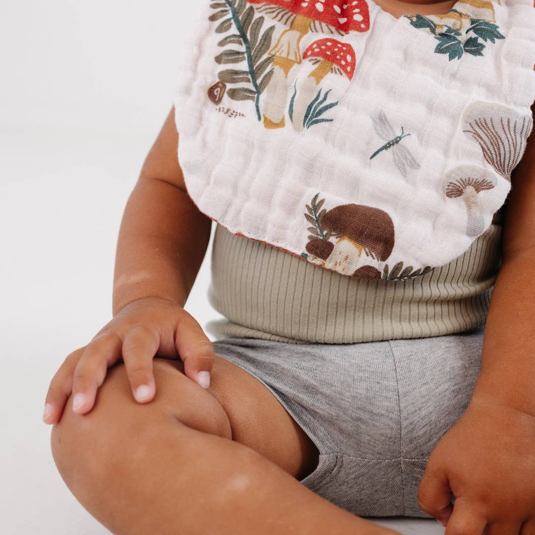 A close-up of a baby wearing a white muslin bib with a colorful forest print of mushrooms, leaves, and dragonflies, paired with a ribbed sage green top and gray shorts.