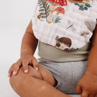 A close-up of a baby wearing a white muslin bib with a colorful forest print of mushrooms, leaves, and dragonflies, paired with a ribbed sage green top and gray shorts.