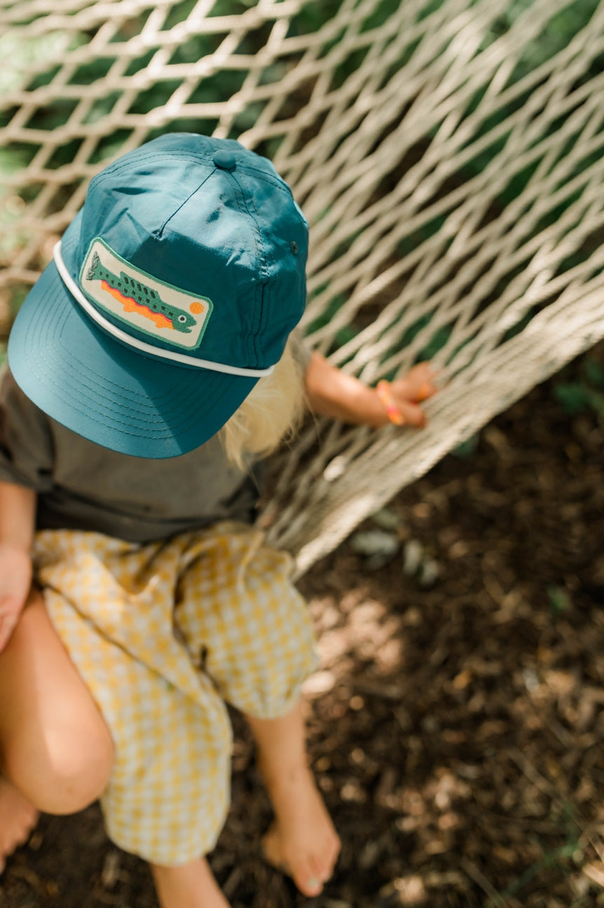 A child sitting in a rope hammock outdoors, wearing a teal baseball cap with a fish patch and yellow-and-white gingham shorts.
