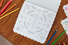 A white scalloped children's coloring book or activity sheet with the number 1 and the word UNO, photographed on a wooden table with colored pencils around it.