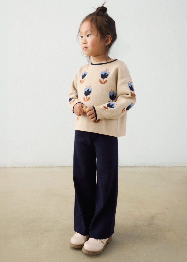 Child wearing a beige sweater with floral patterns and dark pants on a plain background