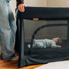 A beige UPPAbaby Remi V2 Playard set up indoors with a baby lying inside, while an adult stands next to it.