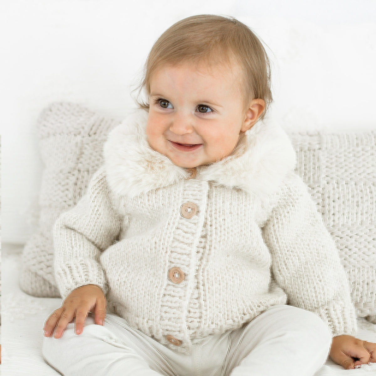 A baby wearing a cream-colored chunky knit cardigan with a faux-fur collar and wooden buttons, sitting on a white textured sofa.