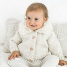 A baby wearing a cream-colored chunky knit cardigan with a faux-fur collar and wooden buttons, sitting on a white textured sofa.