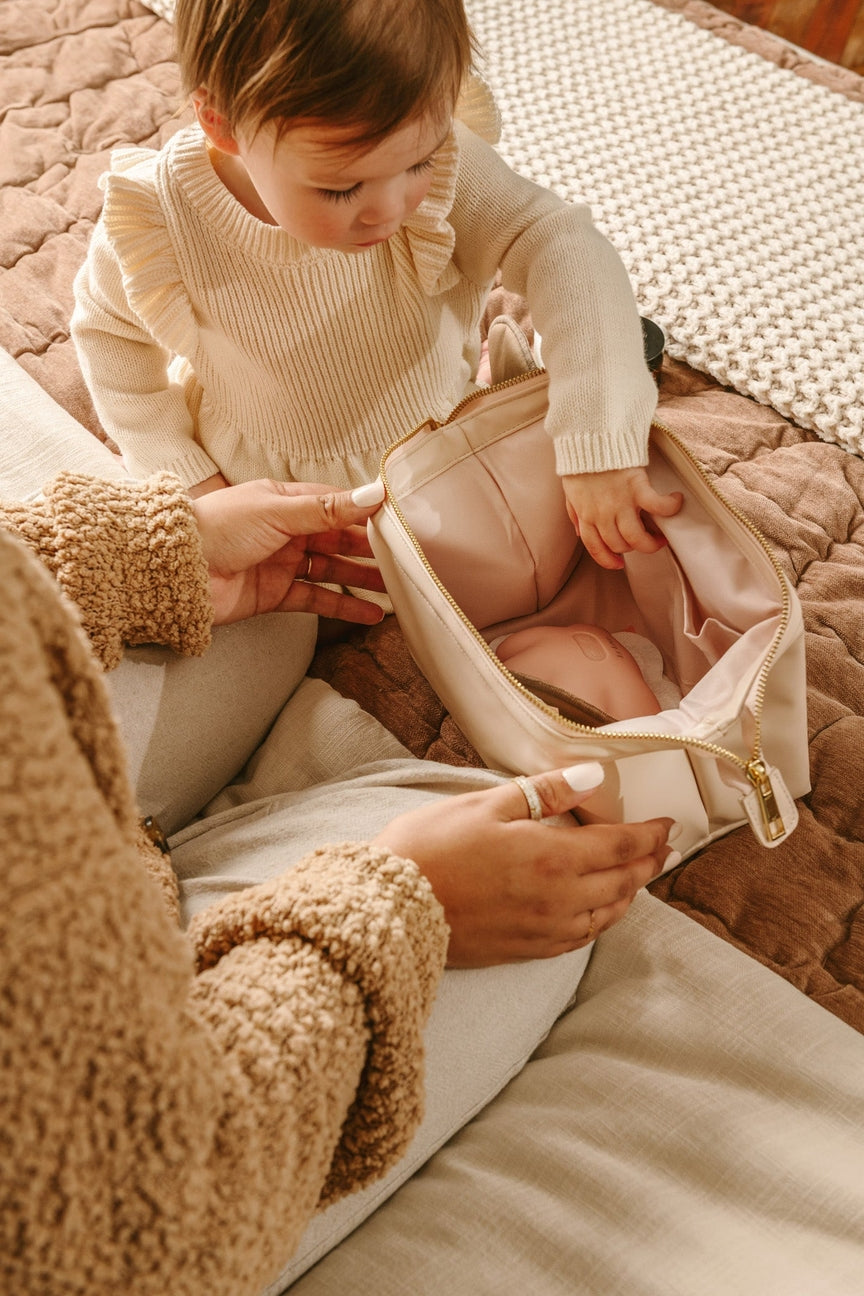 Child in a beige outfit interacting with an adult holding a pink bag on a textured surface.