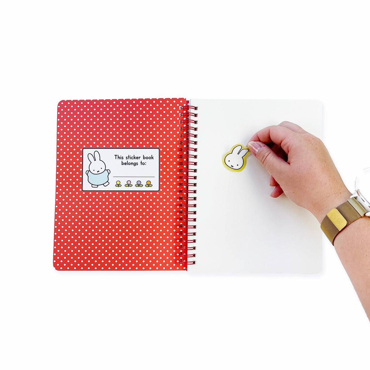 A red and white polka dot spiral-bound sticker book with a white cover page, shown on a white background. A hand is placing a small bunny-shaped sticker on the page.