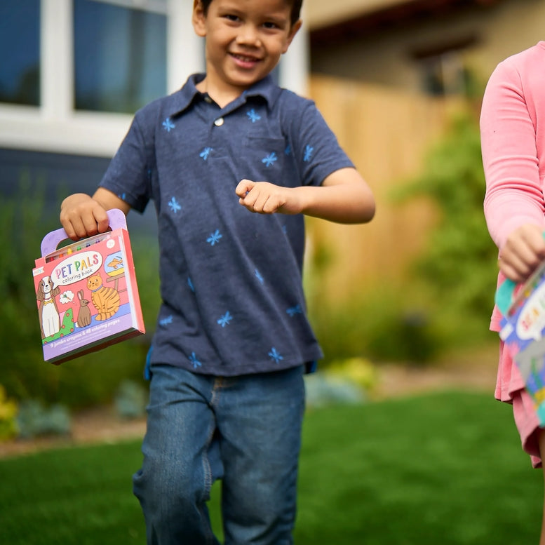 A young boy smiling and running outdoors in a backyard while holding a colorful 'Pet Pals' book box with a purple handle. Another person in a pink shirt is partially visible on the right.