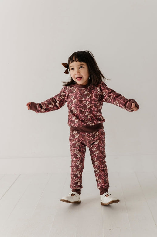 A young child wearing a matching long-sleeve top and jogger pants set with a dusty rose and burgundy floral pattern, standing against a plain light gray studio background.