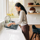 A woman with curly dark hair washes a black sink in a bright, modern kitchen with white countertops and light wood cabinets.