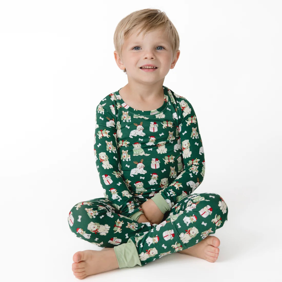 A young child sitting cross-legged on a white studio background, wearing a green long-sleeve pajama set with a festive Christmas print of Santa Claus, snowman, and gift boxes.