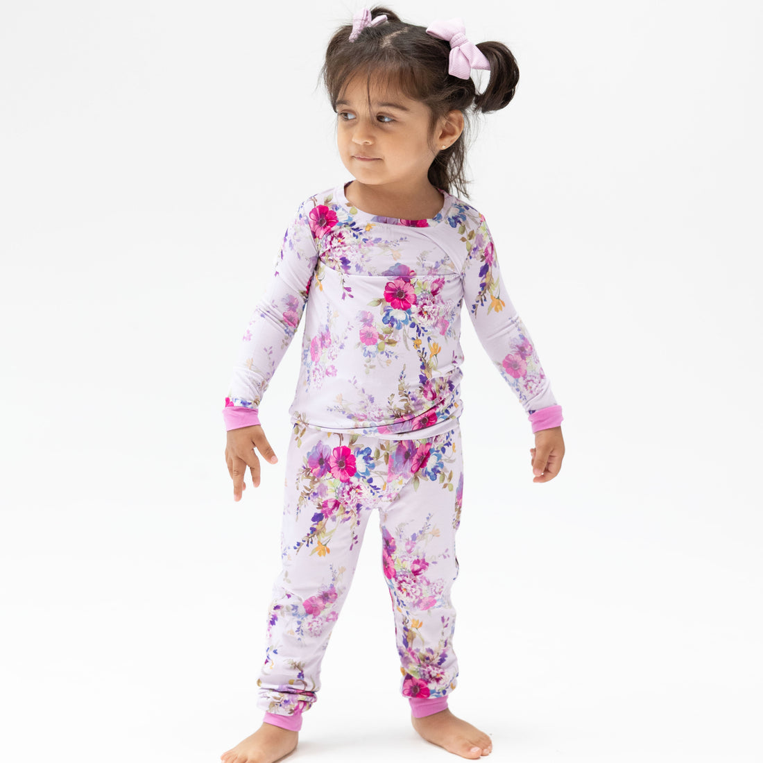 A young girl with her hair in a high ponytail wearing a two-piece pajama set. The pajama set has a light pink base with a colorful floral print in shades of purple, pink, and blue, with solid pink cuffs.