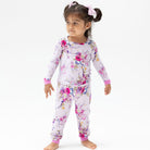 A young girl with her hair in a high ponytail wearing a two-piece pajama set. The pajama set has a light pink base with a colorful floral print in shades of purple, pink, and blue, with solid pink cuffs.