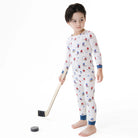 A young boy stands barefoot on a white studio background wearing a long-sleeve pajama set with an all-over hockey player print. He holds a toy hockey stick and puck.