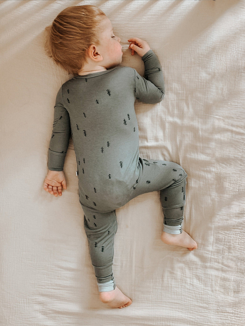 A baby lying on their side on a textured cream-colored bedsheet, wearing a sage green long-sleeve pajama romper with a small black pine tree pattern.