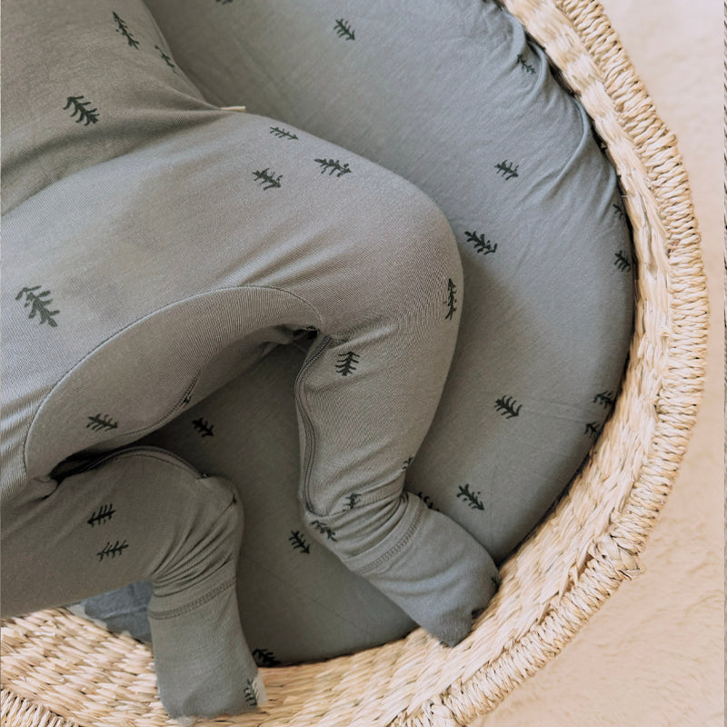 A close-up of a baby wearing soft sage green ribbed footed socks and matching leggings with small black pine tree prints, curled up inside a woven wicker basket.
