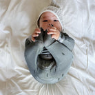 A baby lying on white bedding, wearing a gray knit huggie sleeper with small black tree prints and a matching gray knit hat.