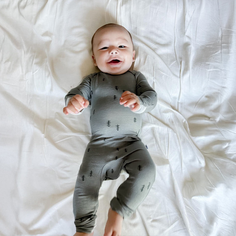 A baby lying on white bedding wearing a gray long-sleeve one-piece jumpsuit with a small dark tree print.