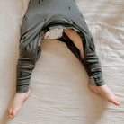 A baby wearing gray harem-style leggings with small black tree prints, lying on a beige textured blanket. The pants are pulled up to show a white diaper.