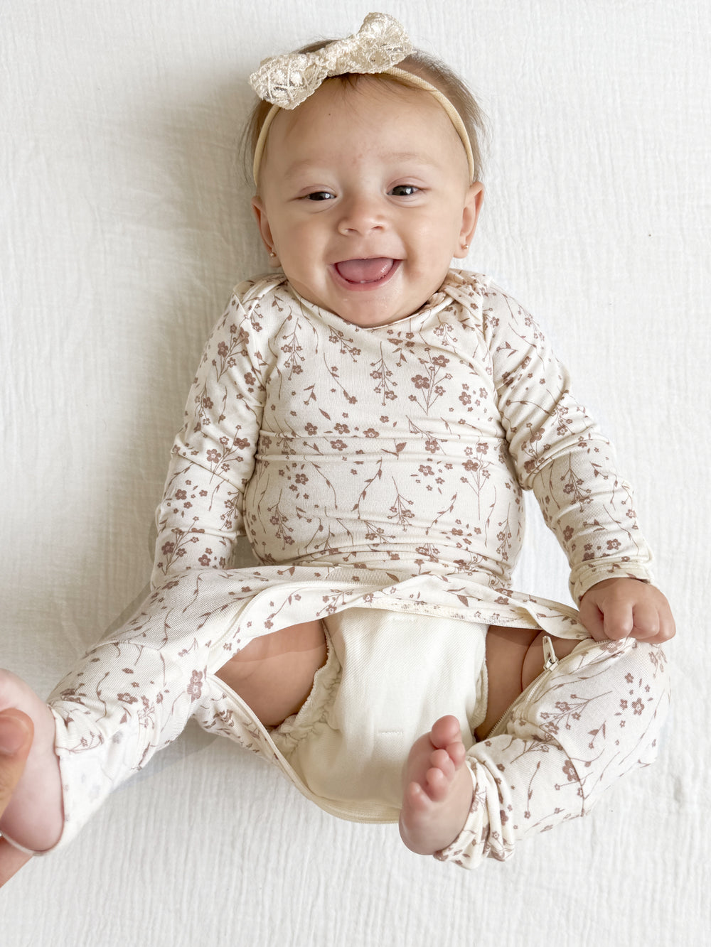 A baby wearing a long-sleeve cream footed romper with a brown floral print and a matching bow headband, lying on a white textured blanket.