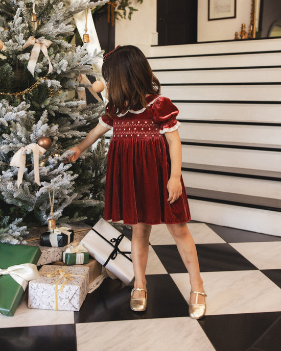 A young girl wearing a festive deep red velvet dress with white lace trim and gold brooch detailing, standing in a holiday-themed room next to a decorated Christmas tree and wrapped gifts.