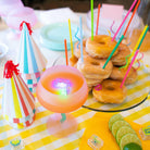 Stack of donuts with colorful straws on a table with party hats and lime slices.