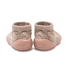 A pair of slip-on knit booties shown from the back, featuring a textured beige cable-knit upper and a dusty pink sole.