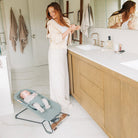 A woman in a bathroom stands beside a baby bouncer with a baby lying inside. The scene includes a wooden vanity, white countertop, towels hanging on hooks, and a mirror reflecting the woman.