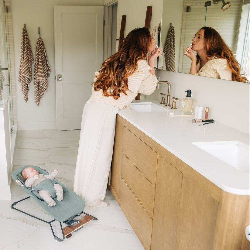 A woman in a white robe stands in a bright bathroom, applying makeup while looking into a mirror. A baby lies on a teal bassinet on the floor nearby.