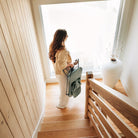 A woman stands on wooden stairs near a large window, holding a sage green folding backpack. The scene is set in a bright, modern interior with light wood walls and a white vase on the landing.