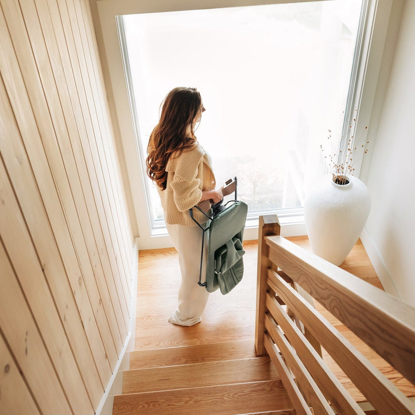 A woman stands on wooden stairs near a large window, holding a sage green folding backpack. The scene is set in a bright, modern interior with light wood walls and a white vase on the landing.