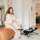 A woman in a cream knit robe sits on a tan leather ottoman in a bright living room, placing a black slipper on a baby monitor scale next to a baby in a black bassinet.