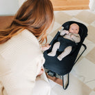A baby in a black baby bouncer chair sits on a checkered tile floor while a woman in a cream ribbed sweater leans in closely.
