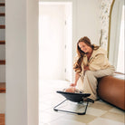 A woman in a cream knit sweater and beige pants sits on a brown leather sofa in a bright, modern room, holding a baby in a black bassinet.