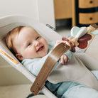 A smiling baby in a light blue outfit sits in a beige baby seat or bouncer, holding a wooden baby teething toy with pastel accents.