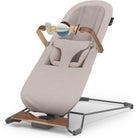 A high-angle studio shot of a baby bouncer chair with a light beige fabric seat, a dark gray metal base, and natural wood accents on the handle and footrest. A wooden toy bar with soft blue, white, and brown rattle-like toys is attached to the handle.