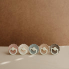 Five colorful baby pacifiers arranged in a row on a beige surface with a brown background.