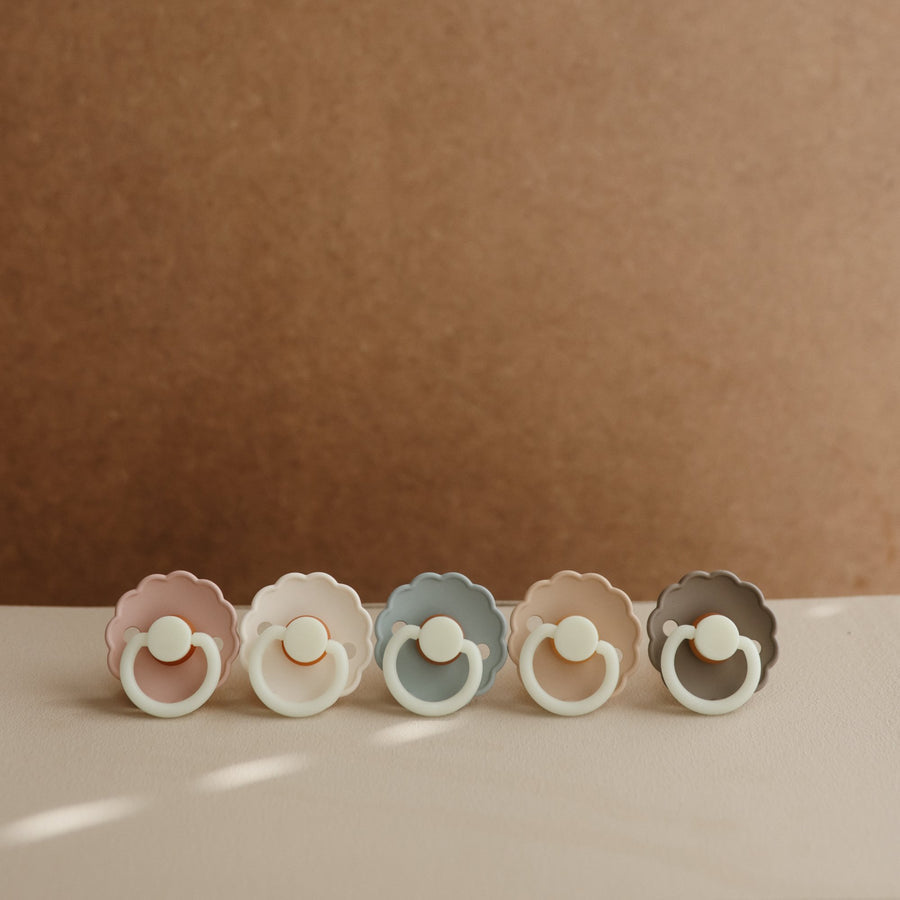 Five colorful baby pacifiers arranged in a row on a beige surface with a brown background.