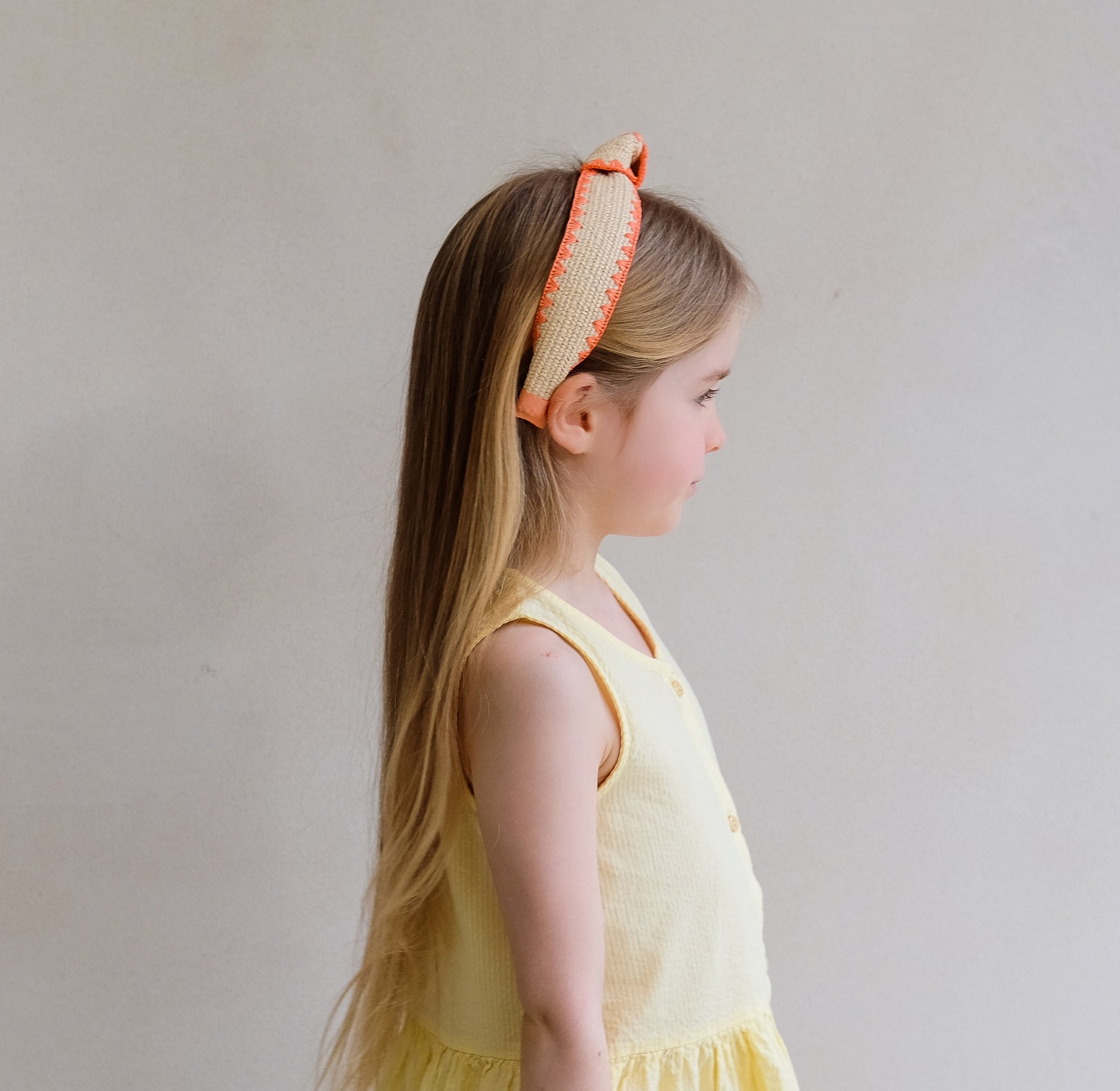 Young girl wearing a yellow dress and an orange headband with a bow on a plain background