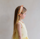 Young girl wearing a yellow dress and an orange headband with a bow on a plain background